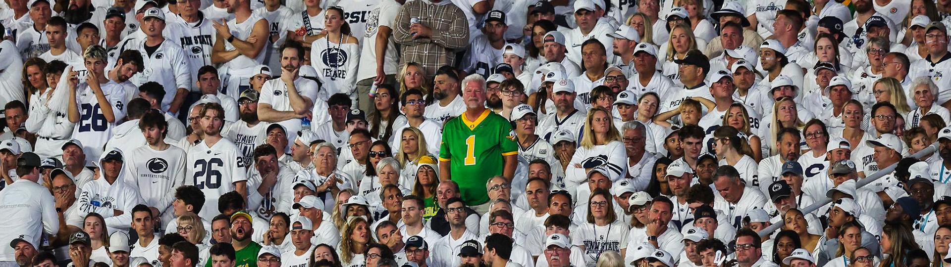 A man in a bright green Oregon jersey stands in the center of a a crowd of Penn State fans dressed in white.