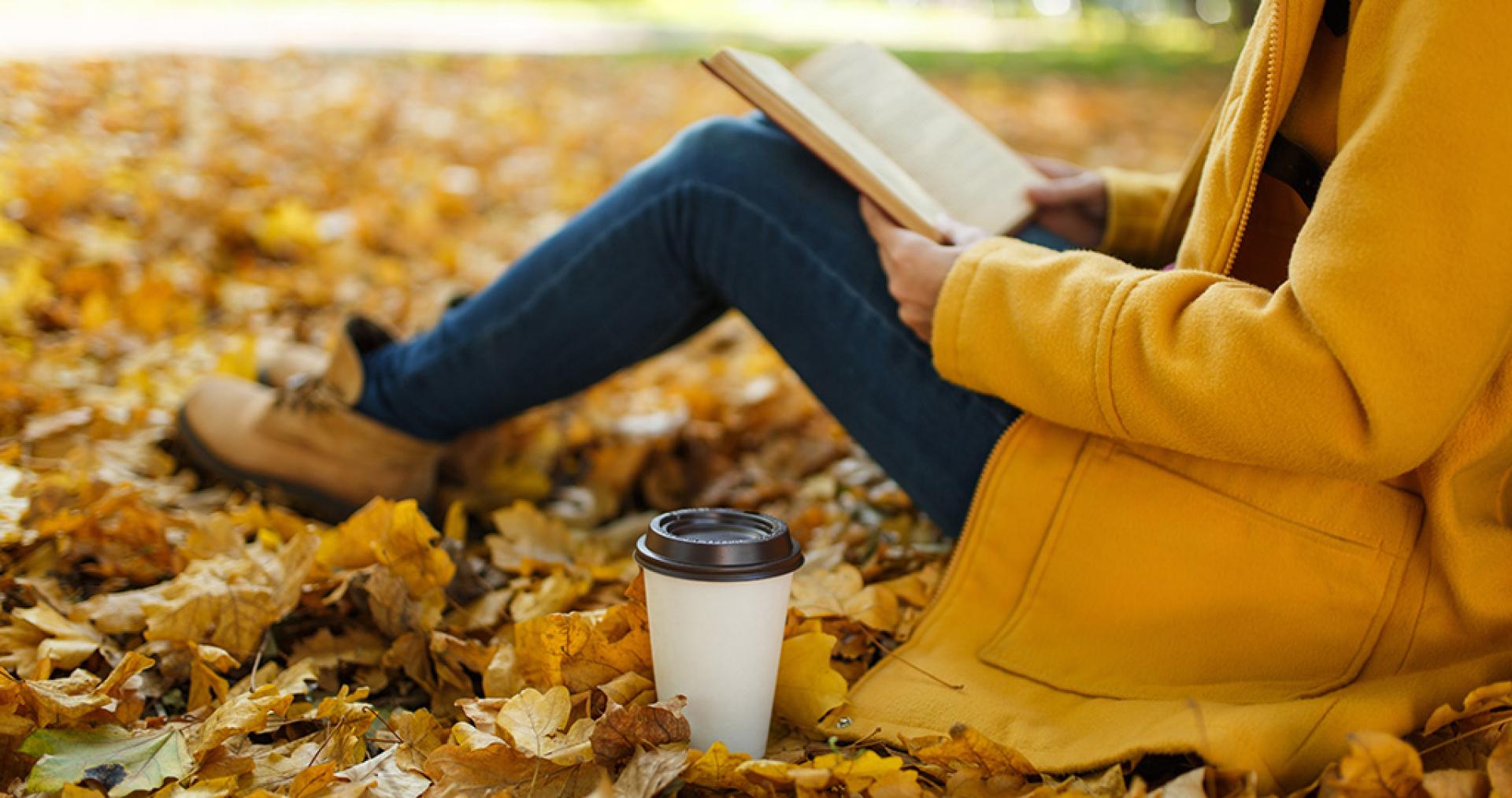 Person in yellow coat reading a book among autumn leaves in a park, with a coffee cup nearby.