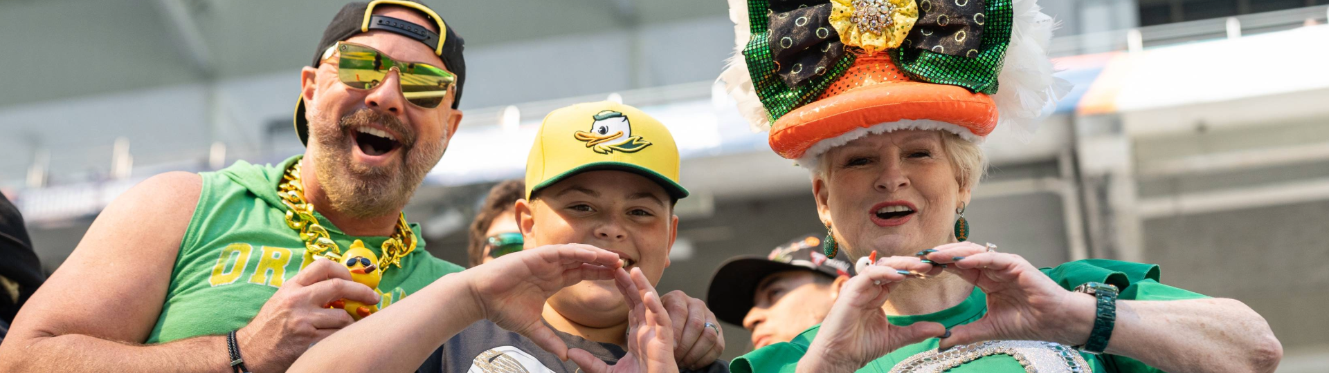 A man in a green Oregon tank top wears a gold chain with a duck on the end, wearing sunglasses. A young boy makes an O with his hands. A middle aged woman wears a sequined hat that looks like an orange duck bill with a green and black bow on top and a fluffy white head on top while making an O with her hands.