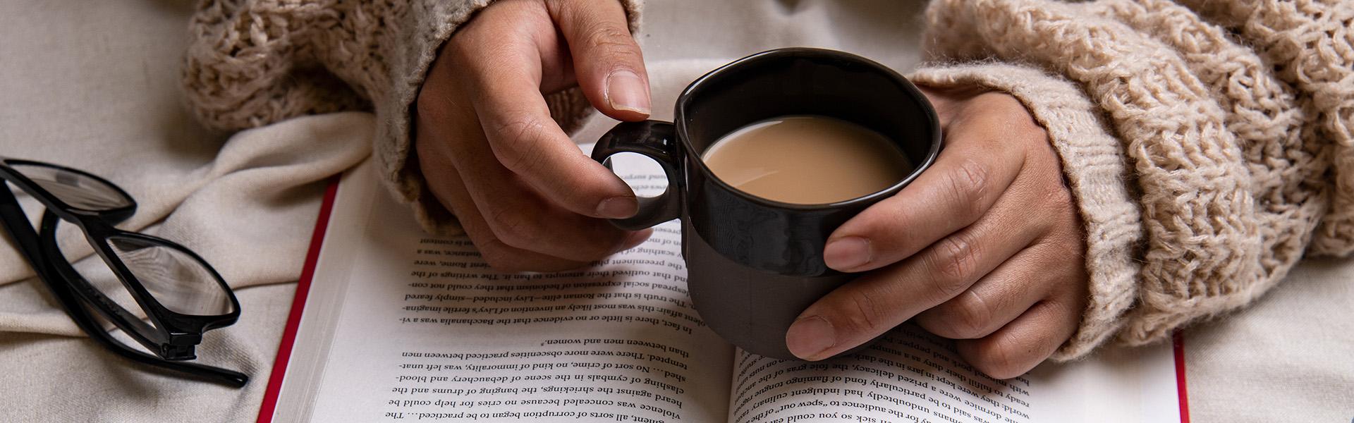 Glasses sit next to an open book. The long sleeves of a tan sweater cover a person's wrists, their hands hold a mug of a warm drink on top of the open pages of the book.