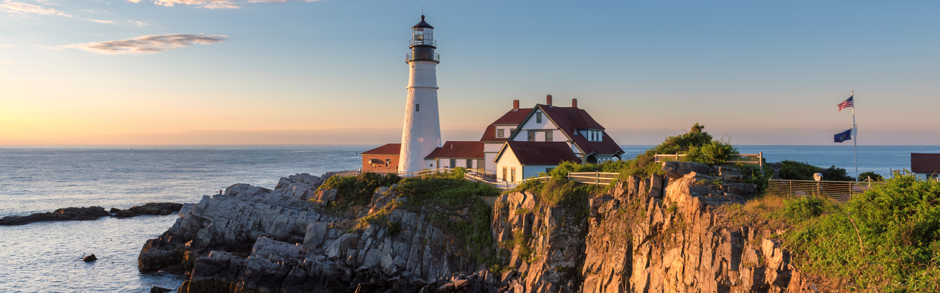 A lighthouse stands on the edge of a cliff above blue waters during sunset.