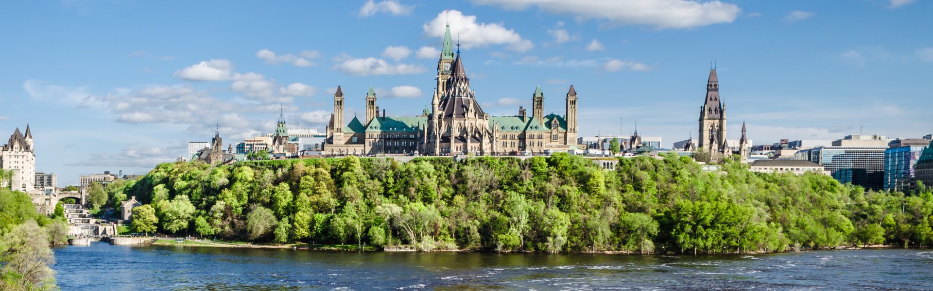 Spires and grand white buildings rise beyond green trees past blue waters.