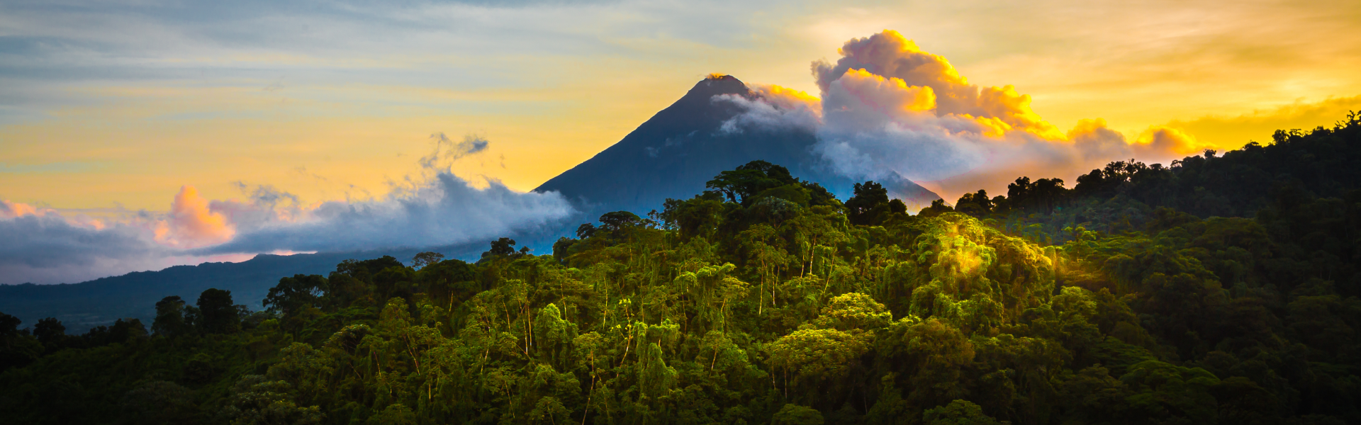 A mountain rises through orange and blue clouds above a lush green forest.