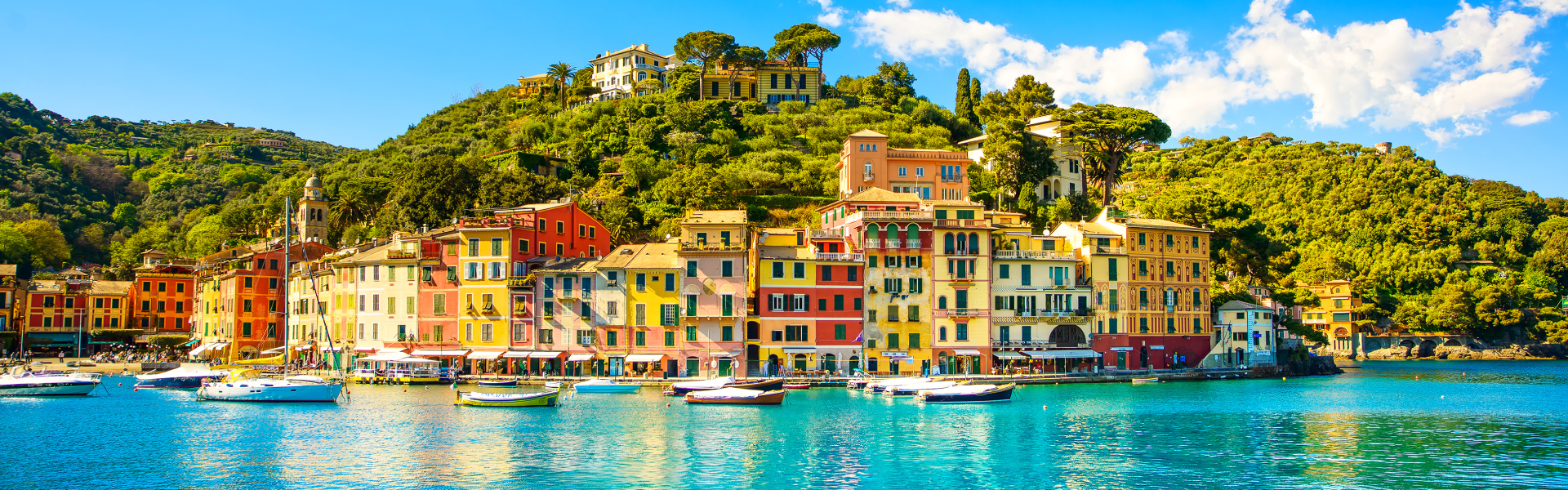 Turquoise waters below multicolored houses on a green tree-covered hillside.