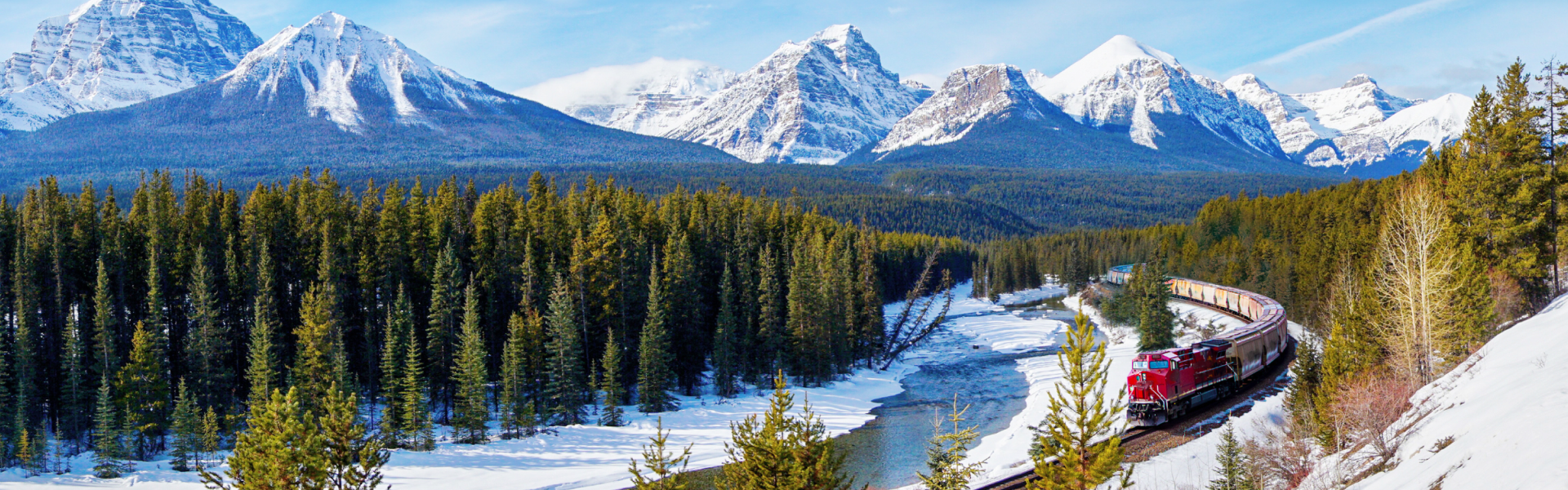 A red train carves between evergreen trees below towering snowy mountains.