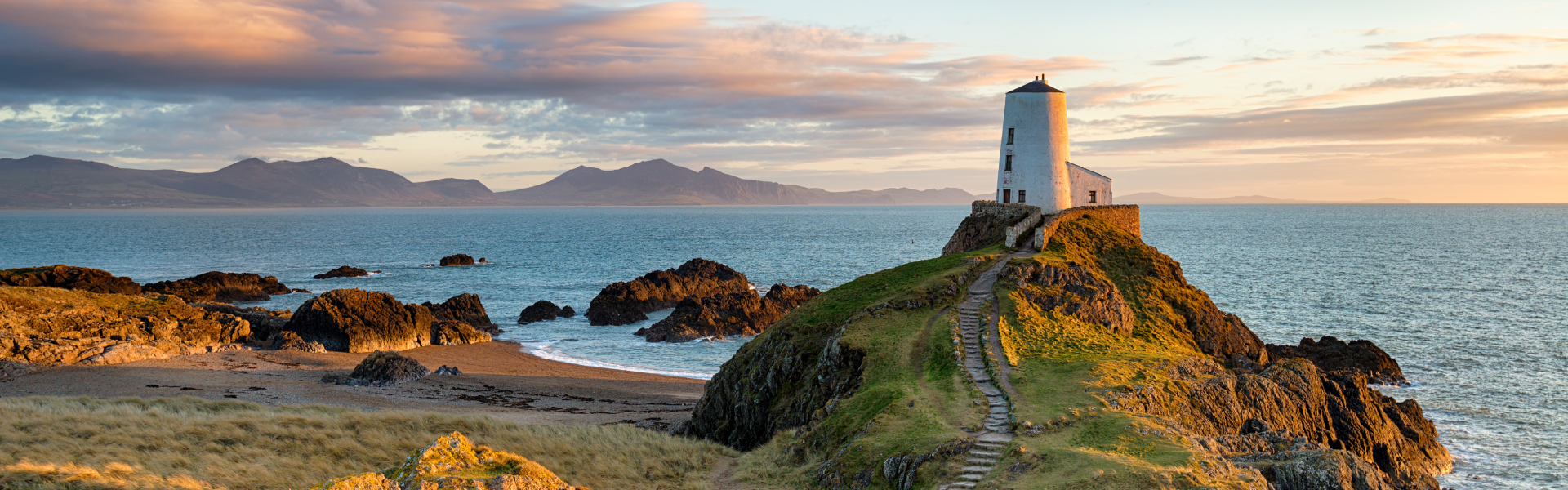 A white stone structure stands on the green edge of a cliff overlooking waters at sunset.