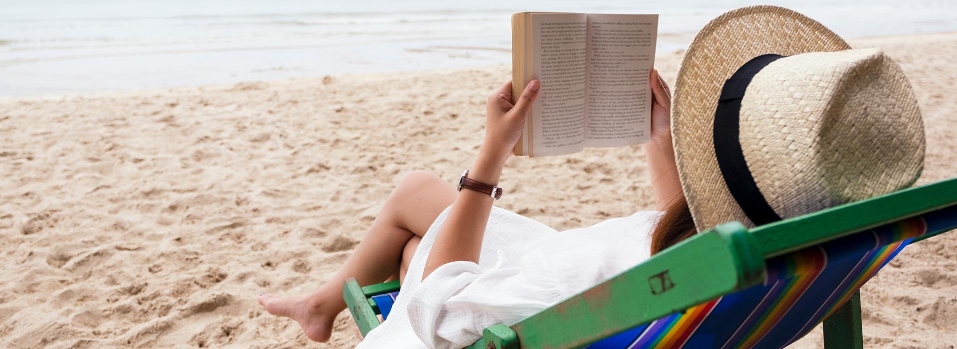 A woman in a straw fedora sits in a green and blue beach chair holding a book with the ocean in the distance.