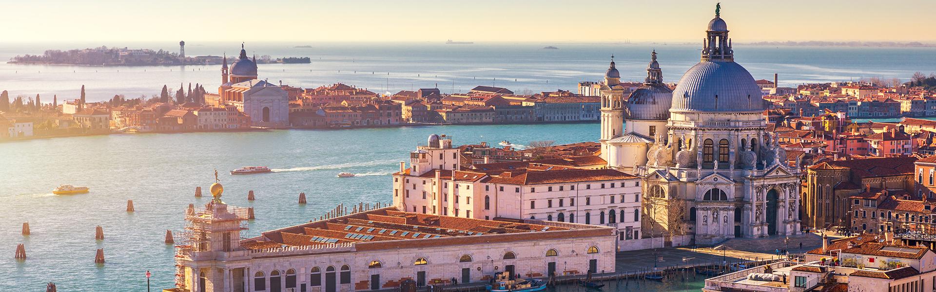 Red-roofed white buildings below a large building with a dome on top along turquoise waters at sunset.