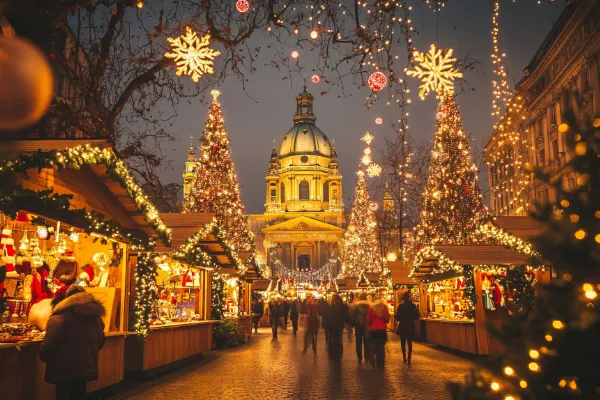 A European Christmas market lane is lit by holiday lights, giant Christmas trees rising into the sky. Vendor stalls glow warmly while people walk the street under snowflake lights and ornaments.