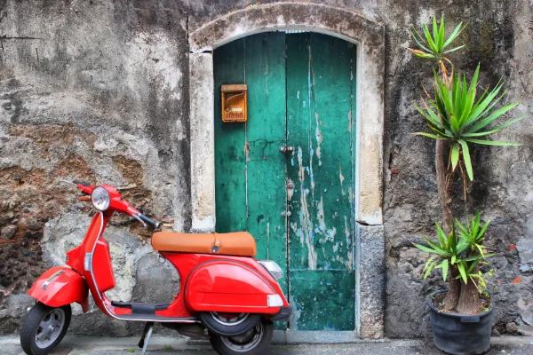A turquoise door stands behind a red Vespa, while a green plant shoots up to the right of the door.
