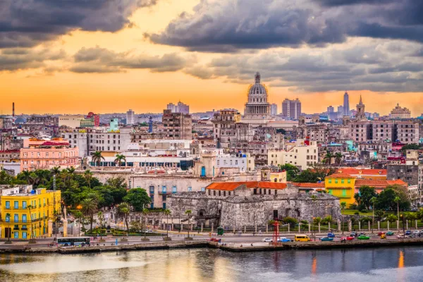 An orange sky and blue clouds hang over a Cuban skyline of colorful buildings.