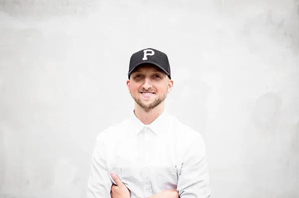 Person in a white shirt and black 'P' cap standing with arms crossed against a plain background.