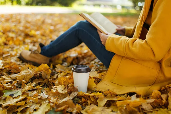Person in yellow coat reading a book among autumn leaves, with a coffee cup nearby.