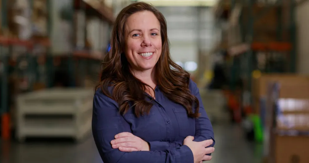 A white female standing in a warehouse smiling with her arms crossed wearing a dark blue shirt. 