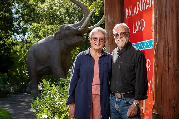 Two people standing by a red ‘Kalapuyan Land’ sign with a large prehistoric animal statue in a garden setting.