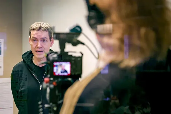 A man stands in front of a camera while he is being interviewed by a person with long hair. 