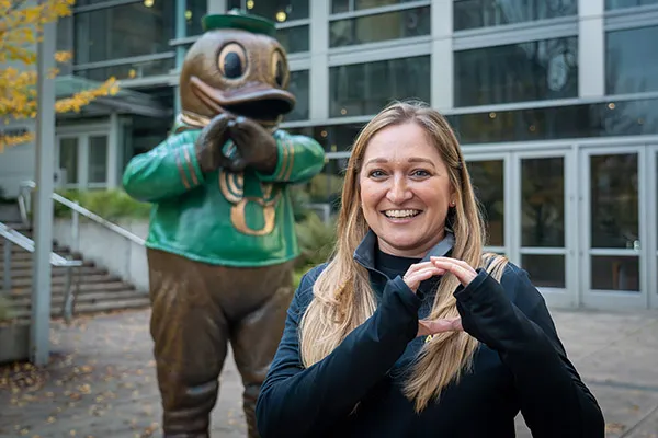 Person making an “O” hand gesture outdoors in front of a large duck statue near a modern building.