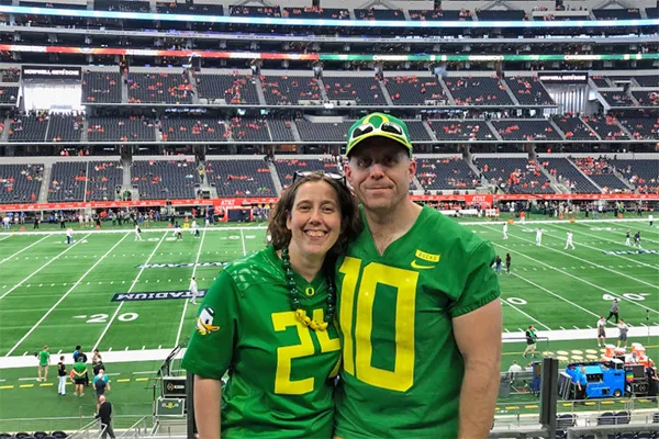 James and Emma Hathaway pose close together wearing bright green Oregon Ducks football jerseys with the inside of a football stadium behind them.