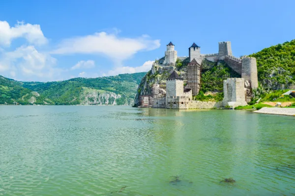 A gray castle on a mountainside sits on the banks of the Danube River's clear blue water, green forested hills and a blue sky stretching behind it.