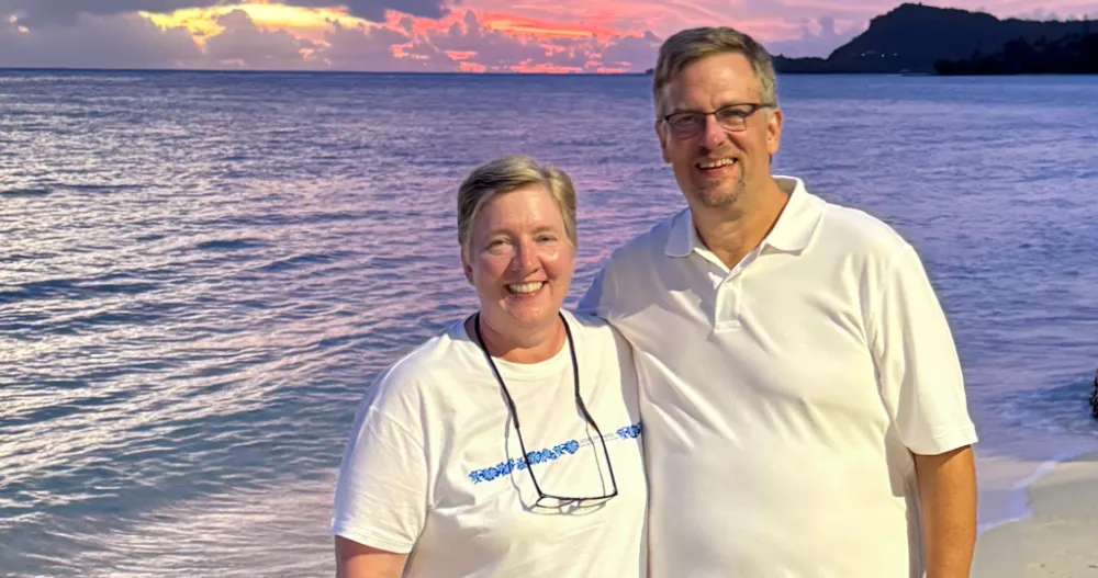 A woman and a man, both in white T-shirts, stand on the beach in front of the ocean.
