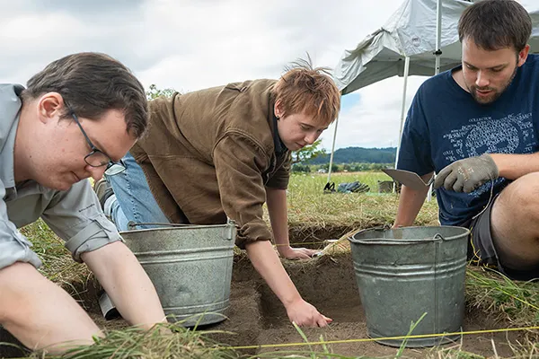 Three young people in work clothes sit on the ground over a square cut out in the dirt with buckets, looking for archeological finds.