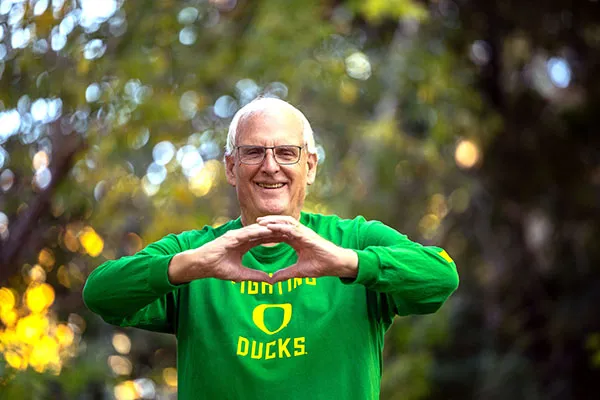 An older white gentleman, Senator Bill Hansell, makes an O with his hands, wearing a bright green long sleeve shirt that says Oregon Ducks in yellow. Blurred trees are behind him.