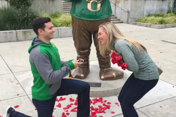 A man kneels on one knee, proposing to a woman in front of the Oregon Duck statue outside the Ford Alumni Center.
