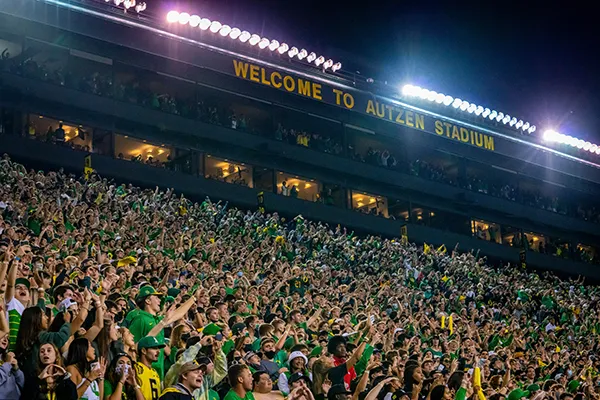 Thousands of people make up a crowd in Autzen Stadium under the glaring lights raising their hands and cheering under a night sky.