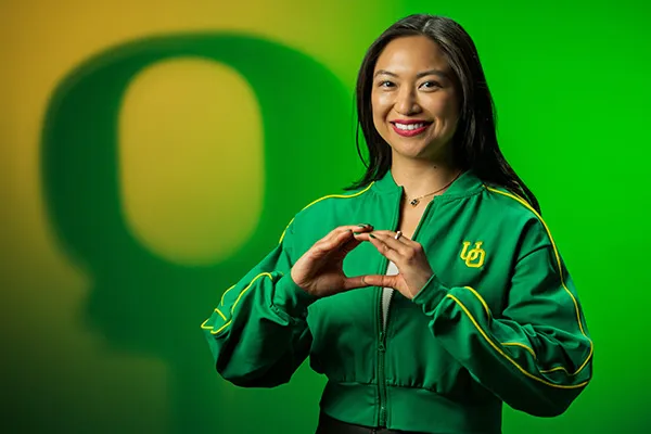 Lorie Acio, wearing a green cropped UO windbreaker, makes an O with her hands in front of a green and yellow lit background with the shadow of an O over her left shoulder.