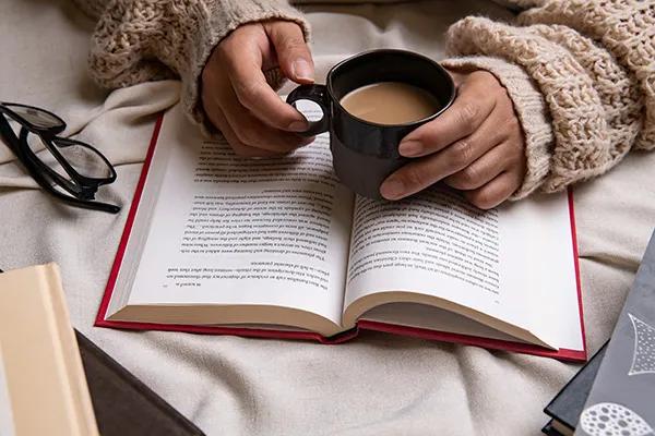 Glasses sit next to an open book. The long sleeves of a tan sweater cover a person's wrists, their hands hold a mug of a warm drink on top of the open pages of the book.