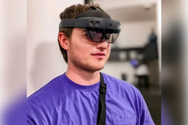 A man in a purple shirt wears a headpiece with dark glasses-like lenses and a band around the head.