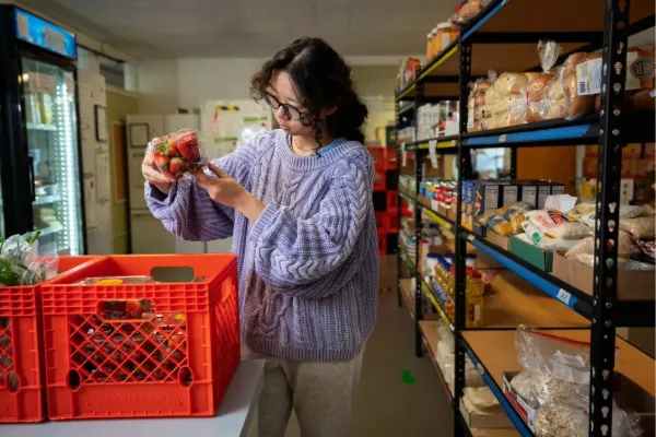 An Asian woman stands next to shelves of food, including rice and bread. She holds a package of strawberries, inspecting it, above a red crate of them. 