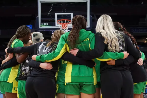 The Oregon Women's Basketball Team face away from the camera toward the net, wrapping their arms around the waists of the women next to them. They are in green uniforms with yellow lines down the sides.