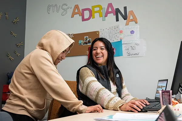 A Latina woman smiles at a desk with a student in a hoodie. The name Ms. Adriana in colorful paper letters is on the wall behind them. 