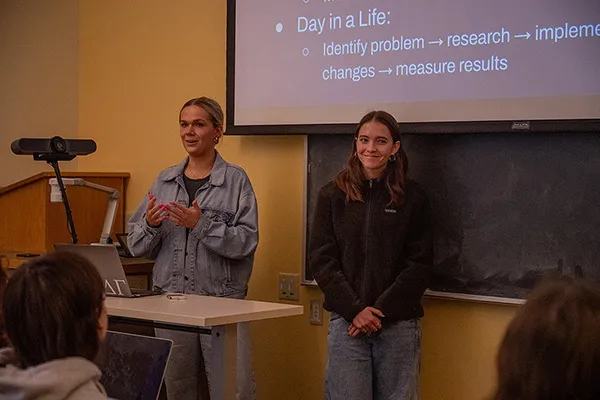 Two young women stand in front of a chalkboard and pull down projector screen presenting in a classroom.