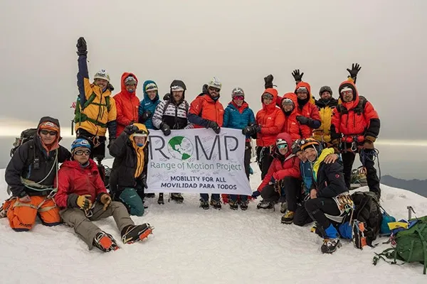 A group of people in climbing gear hold a banner that says ROMP at the summit of Mount Cotopaxi in Ecuador.