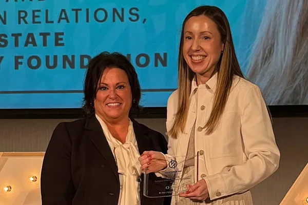 Two women stand together in business clothing. One holds a glass plaque.