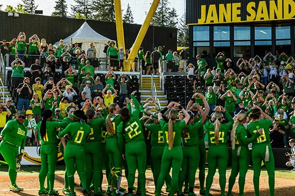 Women in bright green softball uniforms face the audience in the stands, making Os with their hands.