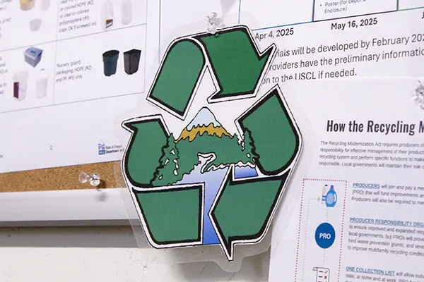 A recycling arrow triangle with a mountain and river in the middle pinned to a cork board in front of papers.