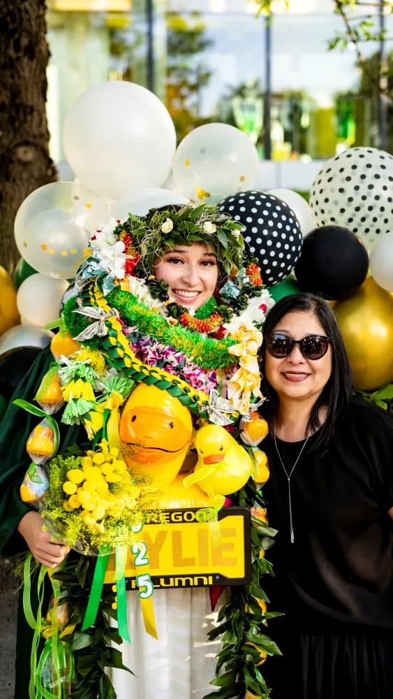 A young woman covered in leis stands next to her mom in front of balloons.