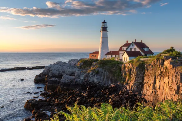 A lighthouse stands on the edge of a cliff above blue waters during sunset.