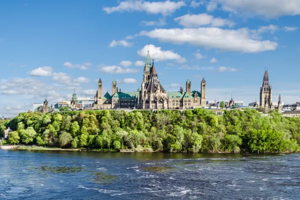 Spires and grand white buildings rise beyond green trees past blue waters.