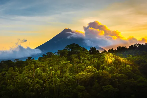 A mountain rises through orange and blue clouds above a lush green forest.