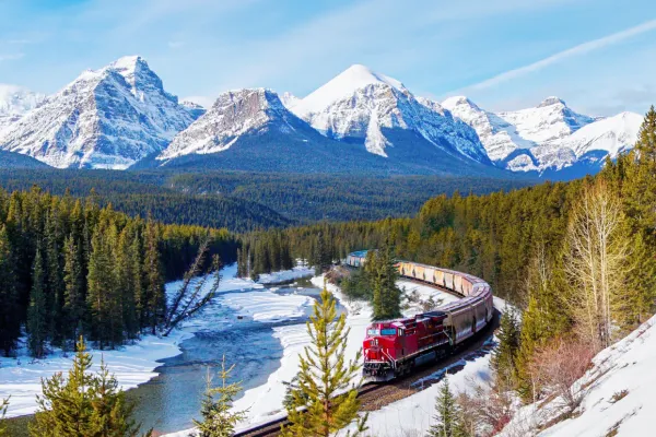 A red train carves between evergreen trees below towering snowy mountains.