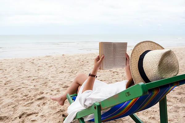 A woman in a straw fedora sits in a green and blue beach chair holding a book with the ocean in the distance.