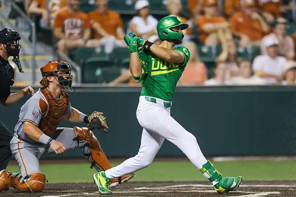 A baseball player in an Oregon Ducks uniform swings a bat with a University of Texas - Austin catcher behind him.