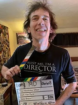 A man holding a film clapperboard smiles at the camera in a shirt that says "Trust me, I'm a director."