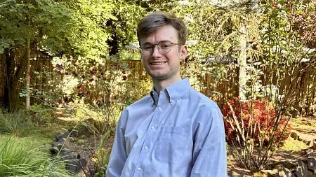 A headshot of a white man in a blue button up shirt in front of trees.