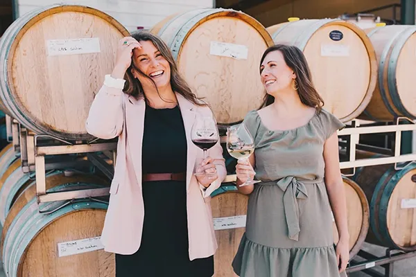 Two women holding wine glasses laugh in front of wine barrels.