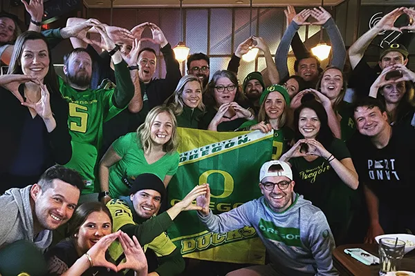 A group of people make Os with their hands in front of a University of Oregon flag.
